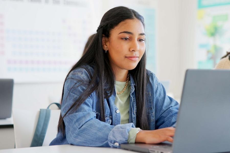 Female college student in a classroom typing on a laptop computer.